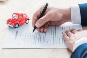 cropped view of car dealer writing in loan agreement near red toy car on wooden tabletop